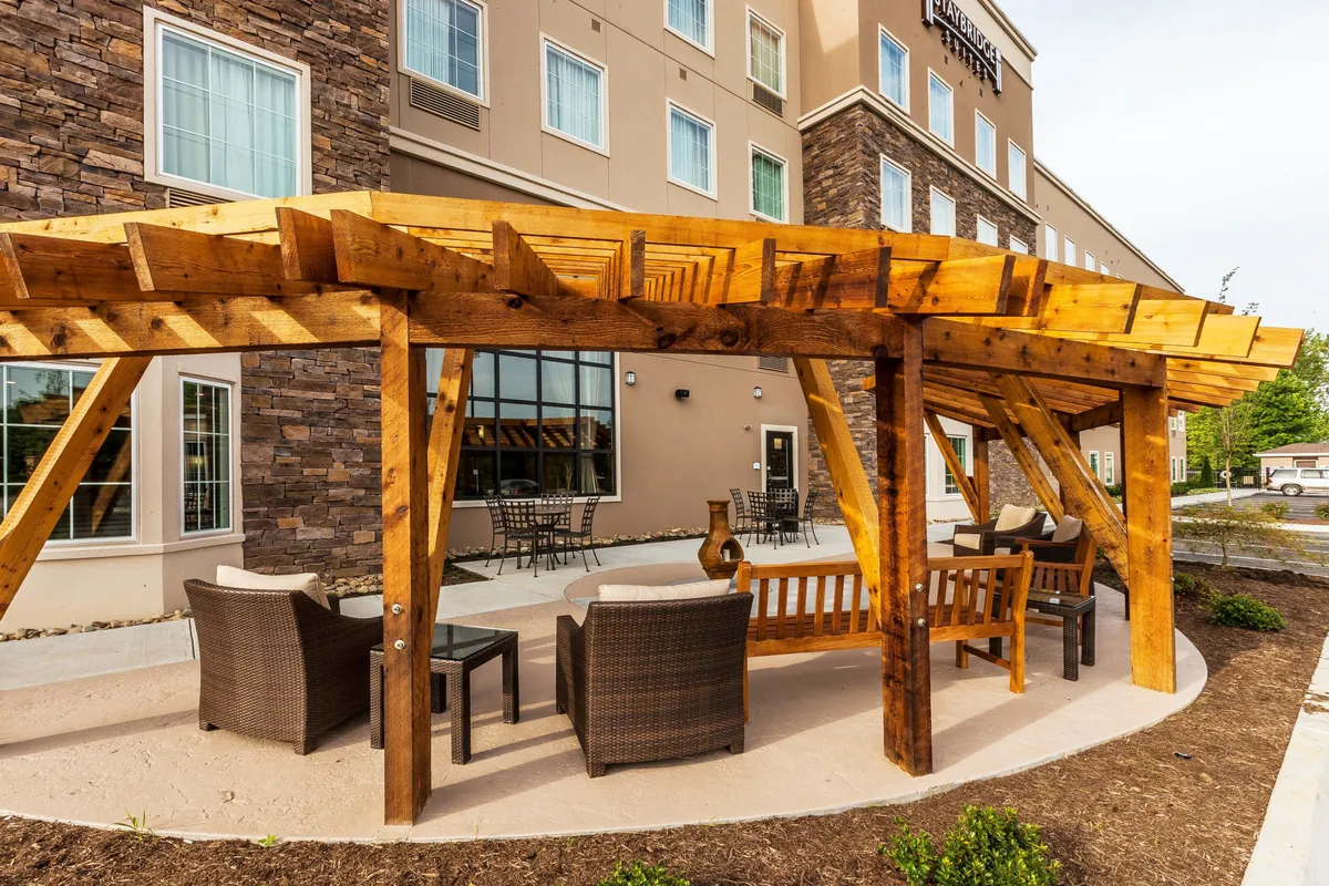 The scene features a wooden pergola with comfortable outdoor seating, stone wall accents, and outdoor tables. The area is bathed in daylight and invites guests to relax, socialize, or enjoy a coffee, blending leisure and casual business interactions in a suburban setting.