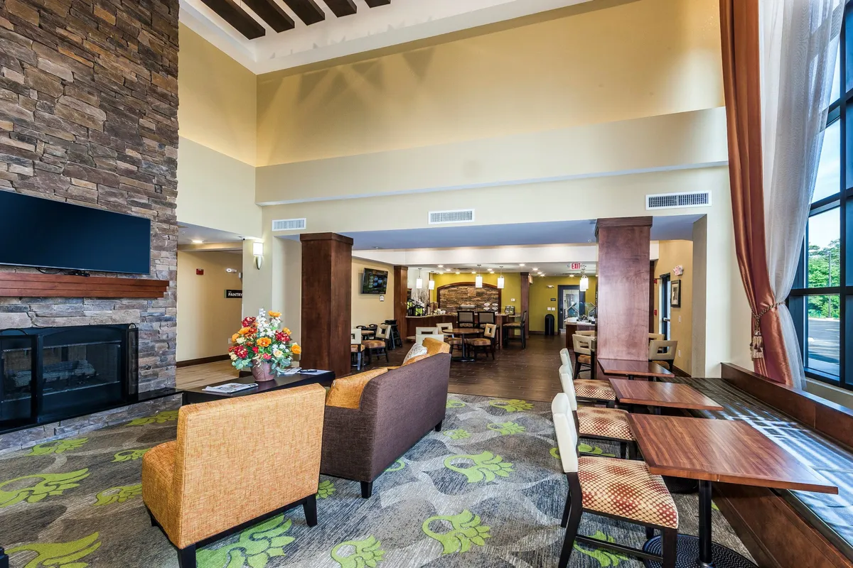 A bright, comfortable seating area featuring a fireplace, large windows that bathe the space in natural light, and a decorative flower arrangement. The arrangement of seating invites relaxation, socializing, or enjoying quiet moments, and the overall mood is cozy yet airy.