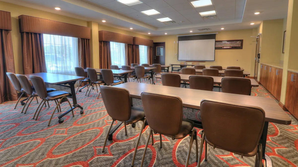 A well-lit interior space with tables and chairs arranged for meetings, featuring large windows that let in abundant natural light. The room is equipped with a projector screen, suggesting flexible uses, and the arrangement conveys a calm, organized atmosphere ideal for quiet work or relaxation.