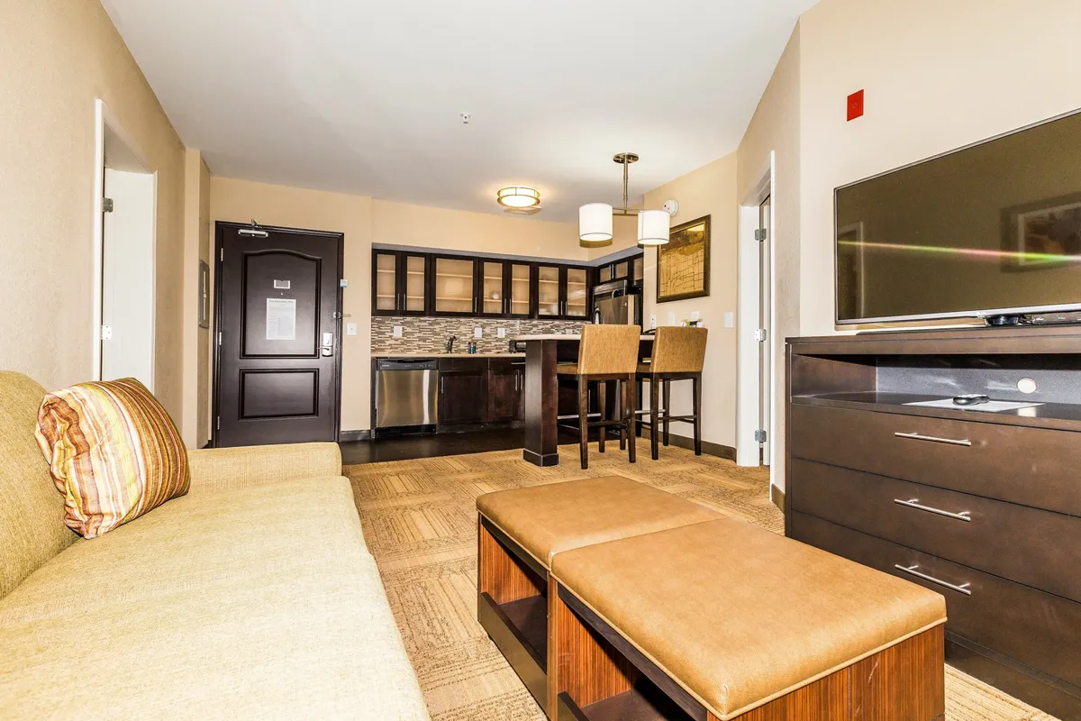 The image showcases a stylish, open-concept living area with a plush sofa adorned with cozy cushions, a sleek kitchen with modern appliances, a dining table set for meals, and a wall-mounted television—all arranged to create a welcoming and functional space.